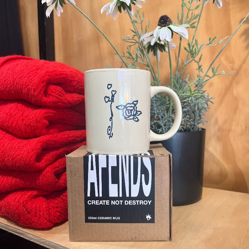 A cream-colored ceramic mug with a black line-art illustration of a flower, displayed on top of its cardboard packaging box. The box is labeled 'AFENDS CREATE NOT DESTROY'.