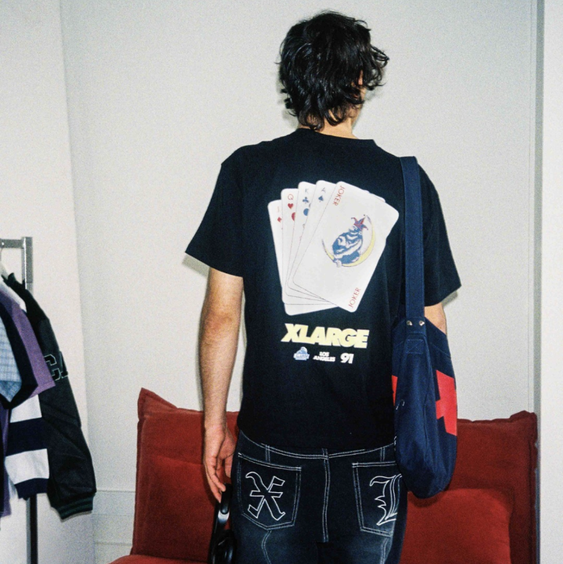 A person standing indoors wearing a black oversized T-shirt with a large white playing-card graphic and the word X-LARGE printed on the back, paired with dark jeans and carrying a blue tote bag. The photo is taken in a room with a red sofa in the background.