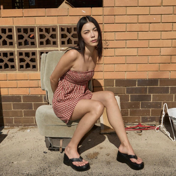 Woman in a red checkered top and skirt sitting on a chair against a brick wall.
