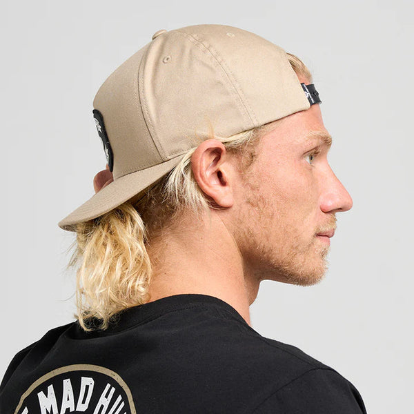 Person wearing a beige cap and black t-shirt with 'The Mad Hueys' logo on a gray background