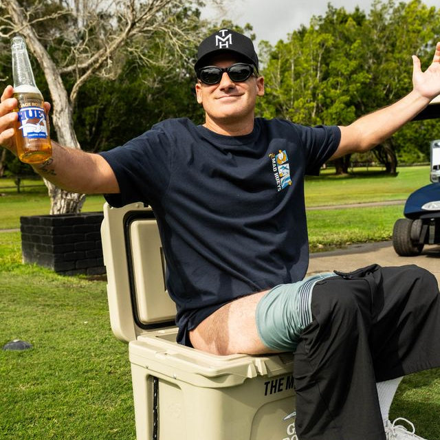 A man sitting on a beige cooler outdoors on a grassy lawn, holding a bottle of beer. He is wearing a navy blue t-shirt with a small graphic on the left chest, black pants, white sneakers, and a black baseball cap.