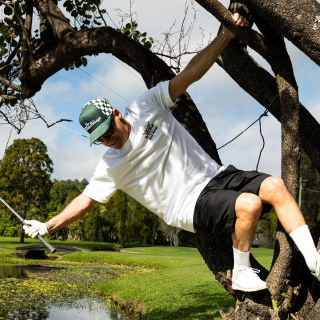 A man in a white t-shirt, black shorts, white socks, and a green cap hangs from a tree branch on a golf course, holding a golf club.