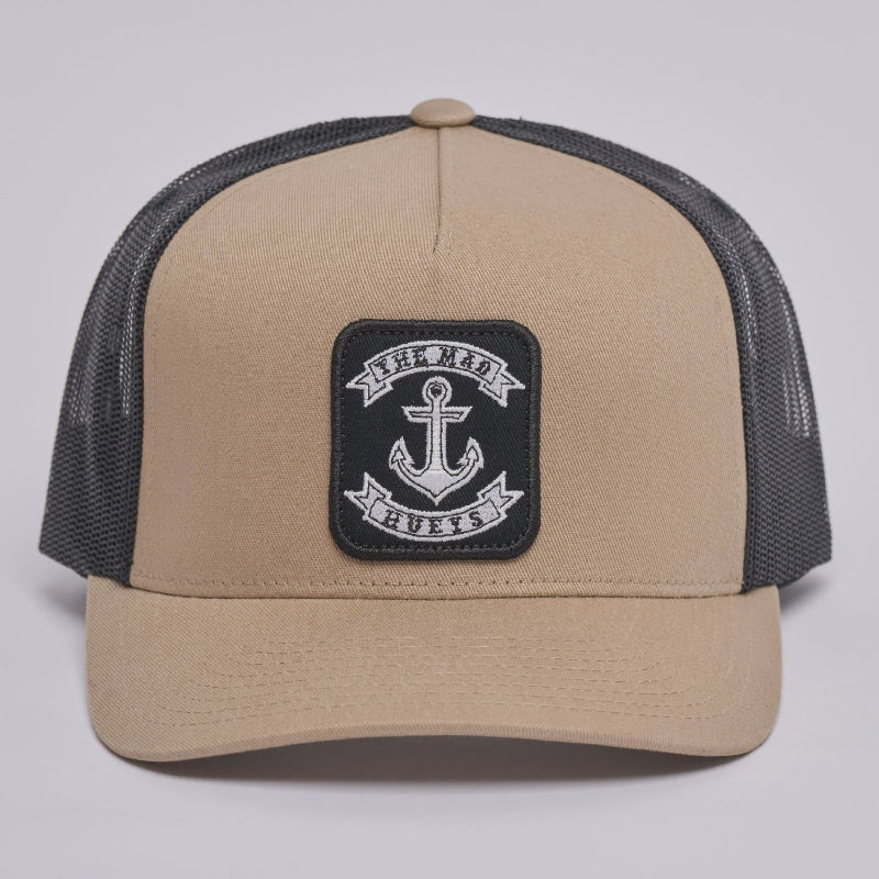 A beige and black trucker hat with a black mesh back and a square black patch on the front featuring a white anchor and the text 'The Mad Hueys'