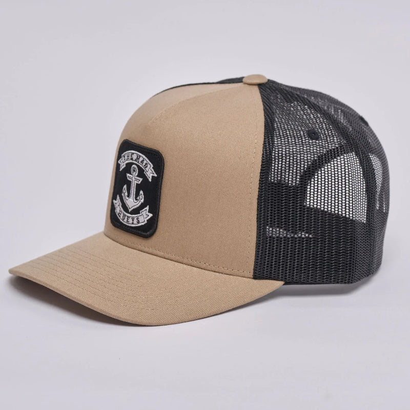 A two-tone trucker cap with a tan canvas front and brim, black mesh back panels, and a black rectangular patch on the front featuring a white anchor logo and the words 'The Mad Hueys'.