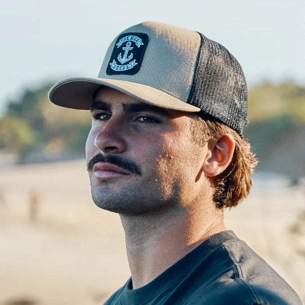 A man wearing a beige and dark gray trucker hat with a black patch featuring an anchor logo and the words 'The Mad Hueys' photographed outdoors on a beach.
