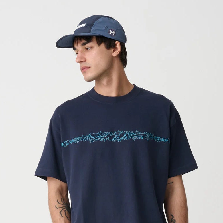 A man modeling a navy blue t-shirt with a blue graphic across the chest, paired with dark gray baggy jeans, black sneakers, and a navy blue baseball cap, against a light gray studio background.