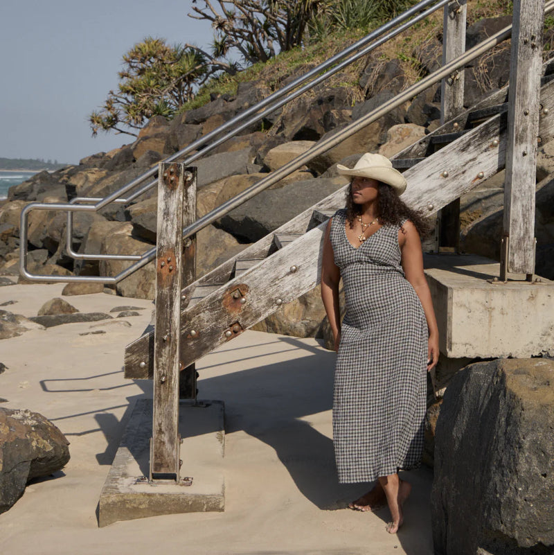 Woman in a checkered dress and hat standing on a rocky beach path with stairs and palm trees in the background.