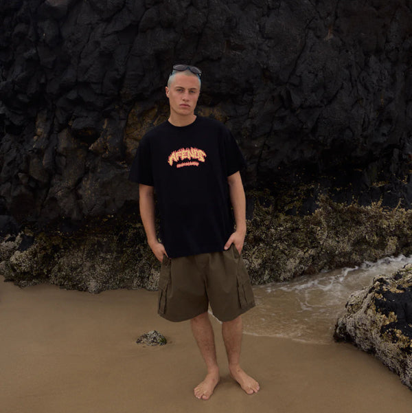 Person wearing a black t-shirt and brown shorts standing on a beach with dark rocky cliffs in the background.