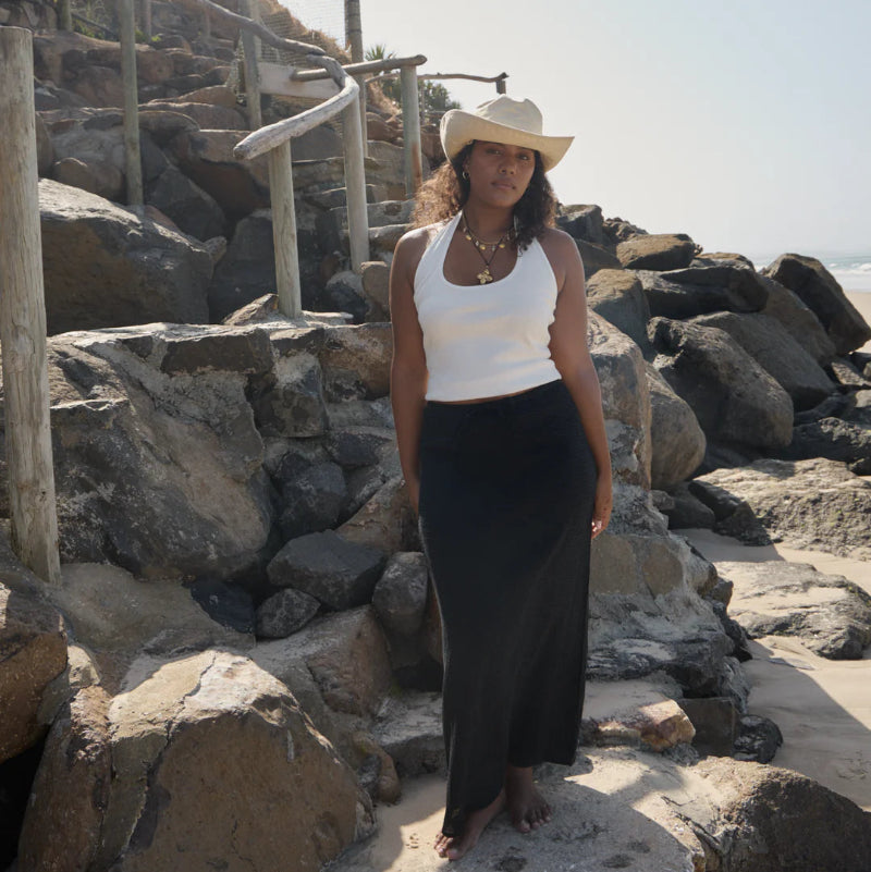 Woman standing on rocks wearing a white tank top and black skirt with a hat.