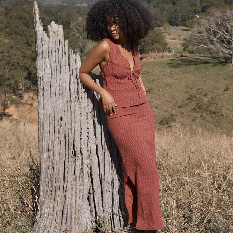 A woman with an afro hairstyle stands outdoors in a grassy field next to a weathered wooden post, wearing a rust-colored sleeveless top and matching maxi skirt d brown boots, with mountains in the backgroundskirt 