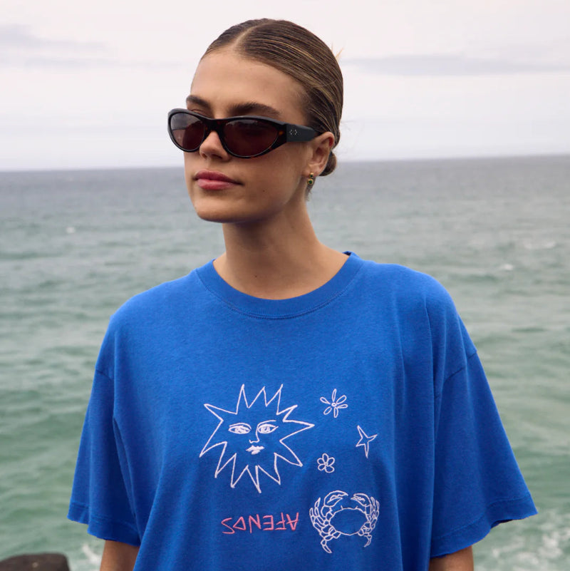 Person wearing a blue t-shirt with graphics and text, standing by the ocean.