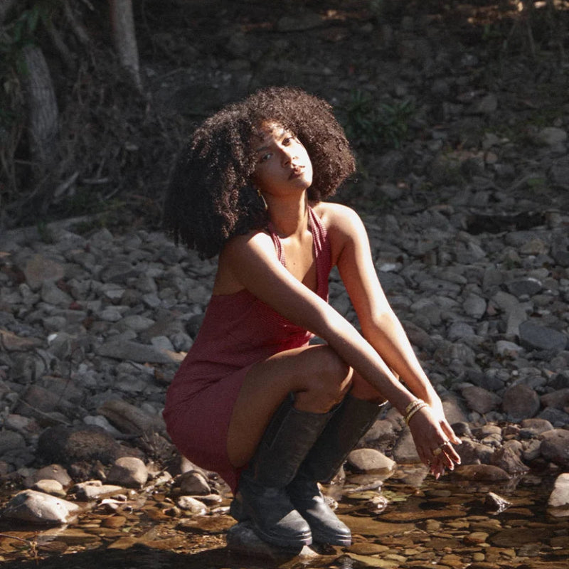 A woman with curly hair crouches in a shallow rocky stream wearing a sleeveless dusty rose dress and dark gray boots, with trees and rocks in the background.