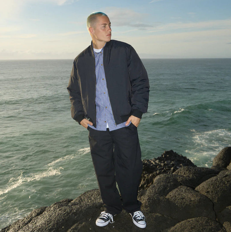 Man standing on rocks by the ocean wearing a black jacket and dark pants.