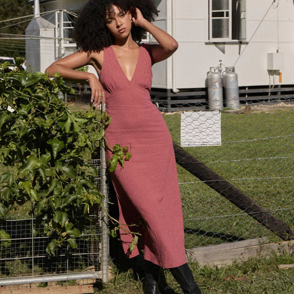 A woman with curly black hair stands outdoors in a grassy yard wearing a dusty rose pink wrap-style midi dress with black ankle boots. A house, fence, and greenery are visible in the background.