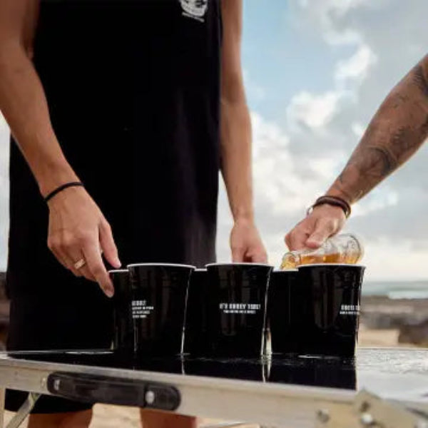 Two people at a beach setting with black cups on a table.