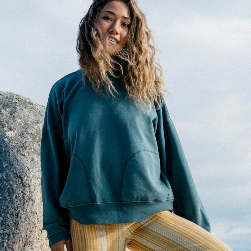 Woman wearing a teal sweatshirt and striped pants standing against a rocky background.