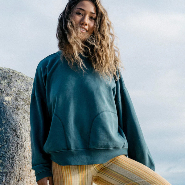 Woman wearing a teal sweatshirt and striped pants standing against a rocky background.