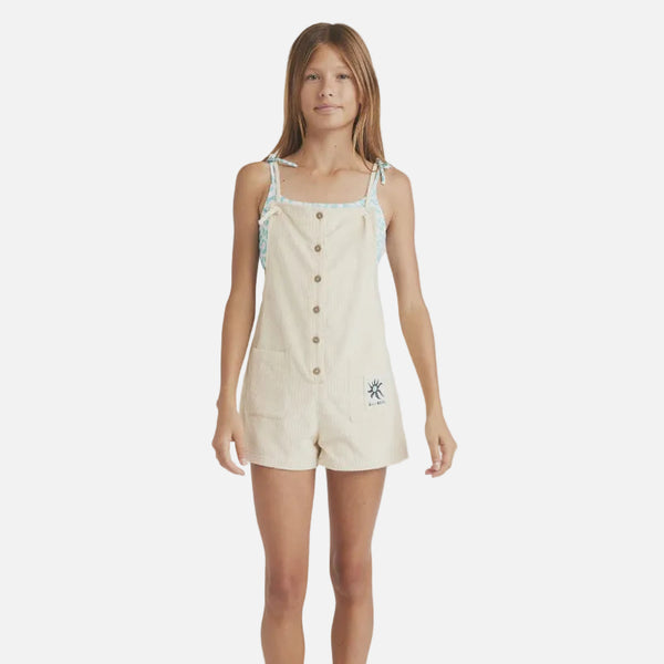 Young girl wearing a beige romper with buttons on a plain background