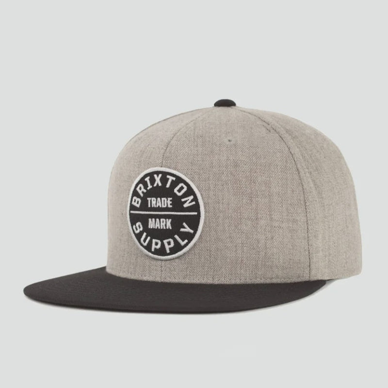 A heather gray snapback cap with a black flat brim and a circular black and white Brixton Supply logo patch on the front, set against a solid light gray background.