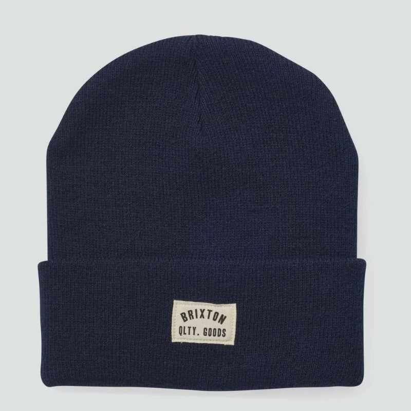 A navy blue knit beanie hat with a folded cuff and a small beige rectangular patch on the front that reads 'Brixton Quality Goods'. The hat is photographed flat against a light gray background.