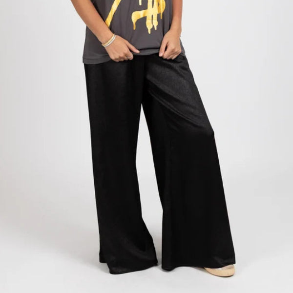 A woman modeling a dark gray t-shirt with a gold heart and dollar sign graphic, paired with black wide-leg pants, against a light gray studio background.