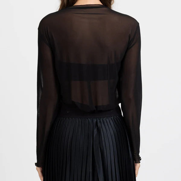 A woman viewed from the back wearing a sheer black long-sleeve top with a horizontal mesh band across the upper back and a black pleated skirt, against a plain light gray studio background.