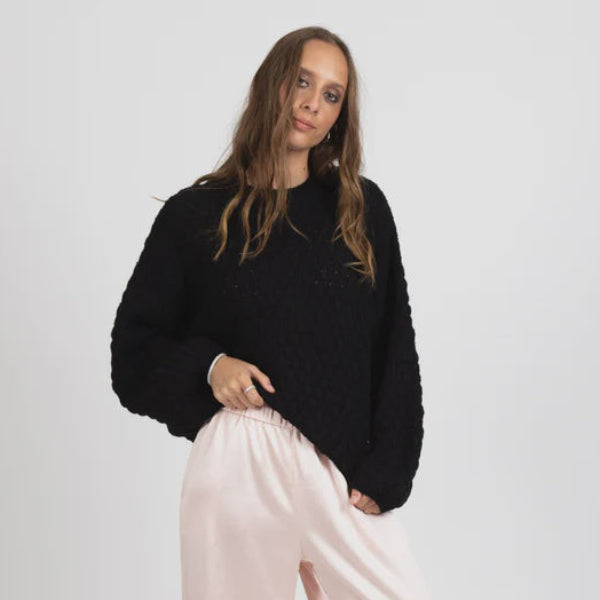 A woman modeling a black textured knit sweater and light pink wide-leg satin pants against a plain light gray studio background.
