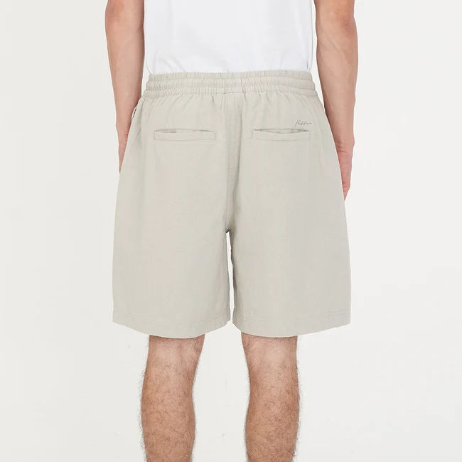 A person shown from the back wearing light beige drawstring shorts, a white t-shirt, white socks, and white sneakers with black accents, against a plain white studio background.