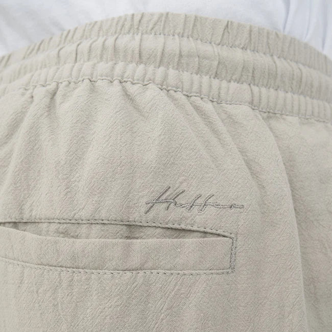Close-up of light beige shorts showing the elastic waistband, a side pocket, and a small embroidered script logo on the fabric.