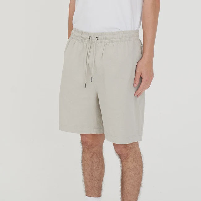 A person wearing light beige drawstring shorts, a white t-shirt, white socks with black branding, and white sneakers, shown from the waist down against a plain white studio background.