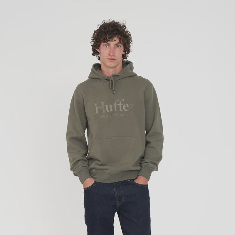 A man wearing an olive green pullover hoodie with 'Huffer' printed on the chest, paired with dark blue jeans and dark sneakers, standing against a light gray studio background.