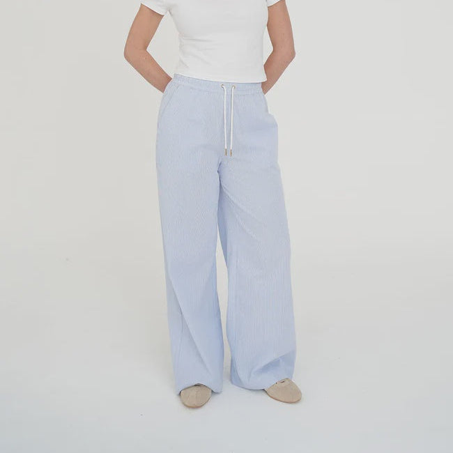 A woman modeling light blue wide-leg trousers with a white drawstring, paired with a white t-shirt and beige shoes, standing against a plain white studio background.