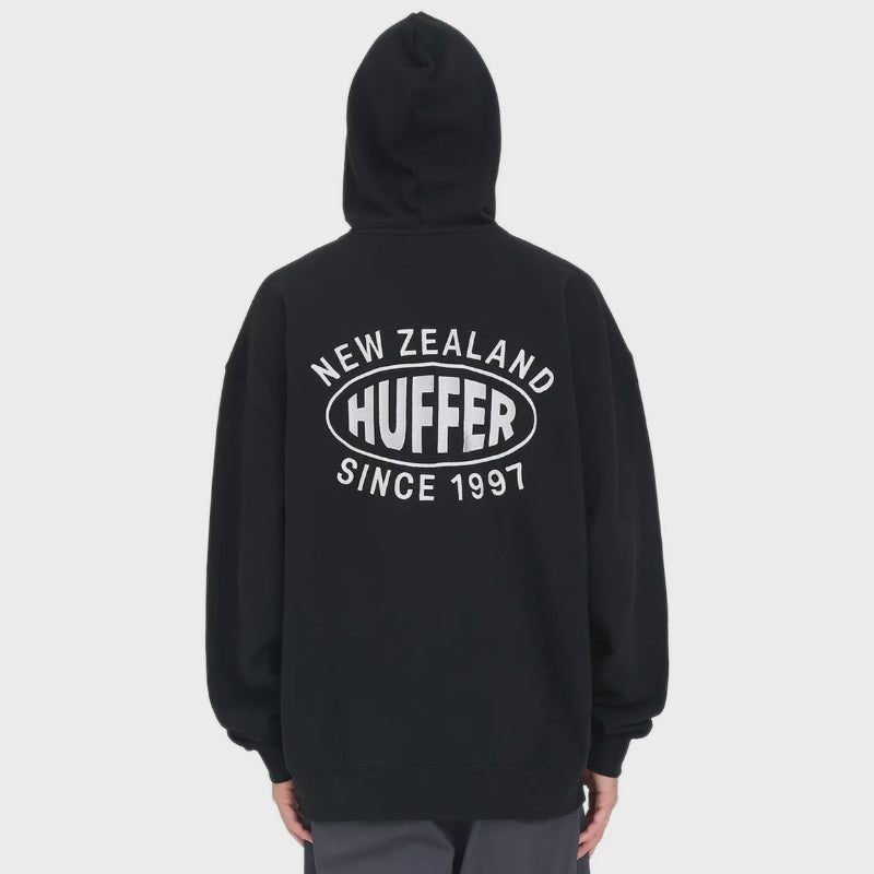 A person shown from the back wearing a black pullover hoodie with a large white graphic on the back reading 'NEW ZEALAND HUFFER SINCE 1997', photographed against a light gray studio background.