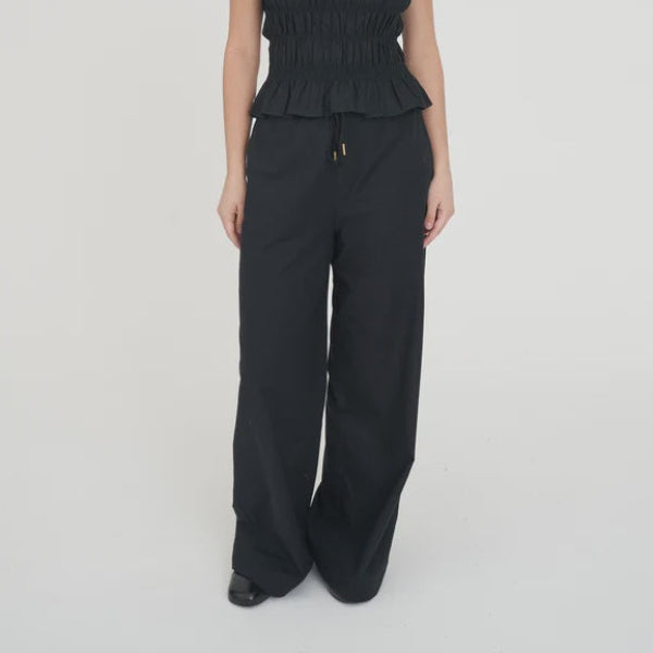 A woman modeling a black sleeveless top with a smocked bodice and ruffled hem, paired with black wide-leg trousers, against a plain white studio background.