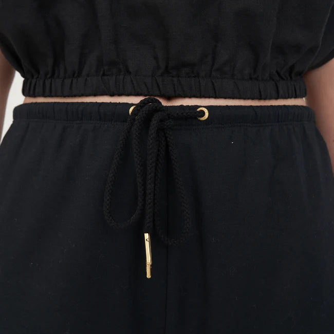 Close-up of a person wearing black drawstring pants with a matching black top, showing the waistband, black drawstring, and small gold-tone hardware.