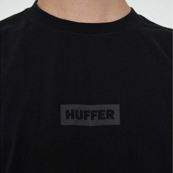 A close-up of a black crewneck t-shirt featuring a tonal dark gray rectangular logo patch on the chest with the brand name 'HUFFER' printed in black.