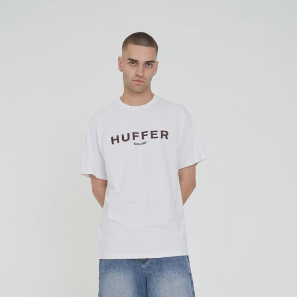 Man wearing a white t-shirt with 'Huffer' logo, blue shorts, and black sneakers on a white background