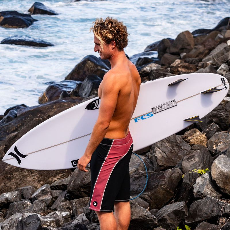A shirtless man standing on a rocky beach holding a white surfboard under his arm, wearing black and pink board shorts, with ocean water and dark rocks in the background.
