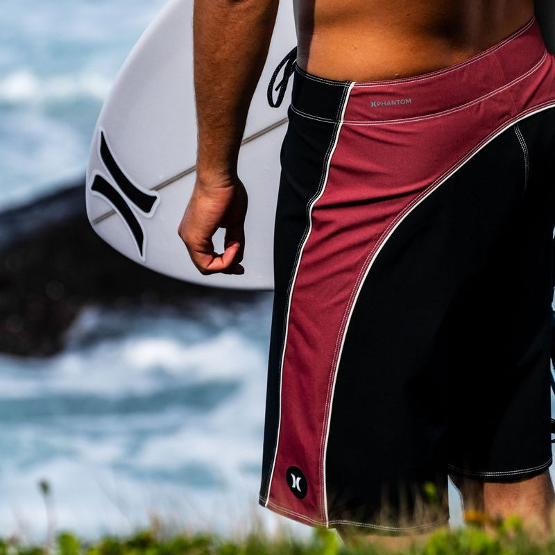 A close-up lifestyle shot of a surfer from behind, wearing red and black boardshorts with white piping and holding a white surfboard. The ocean and rocks are blurred in the background.