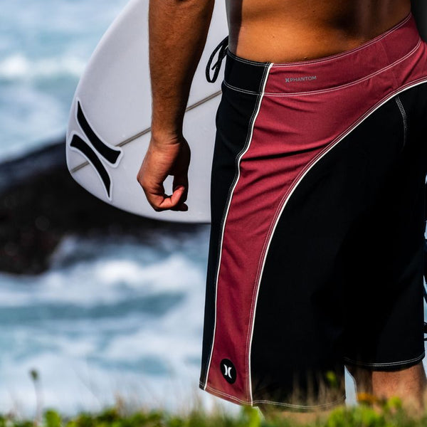 A close-up lifestyle shot of a surfer from behind, wearing red and black boardshorts with white piping and holding a white surfboard. The ocean and rocks are blurred in the background.
