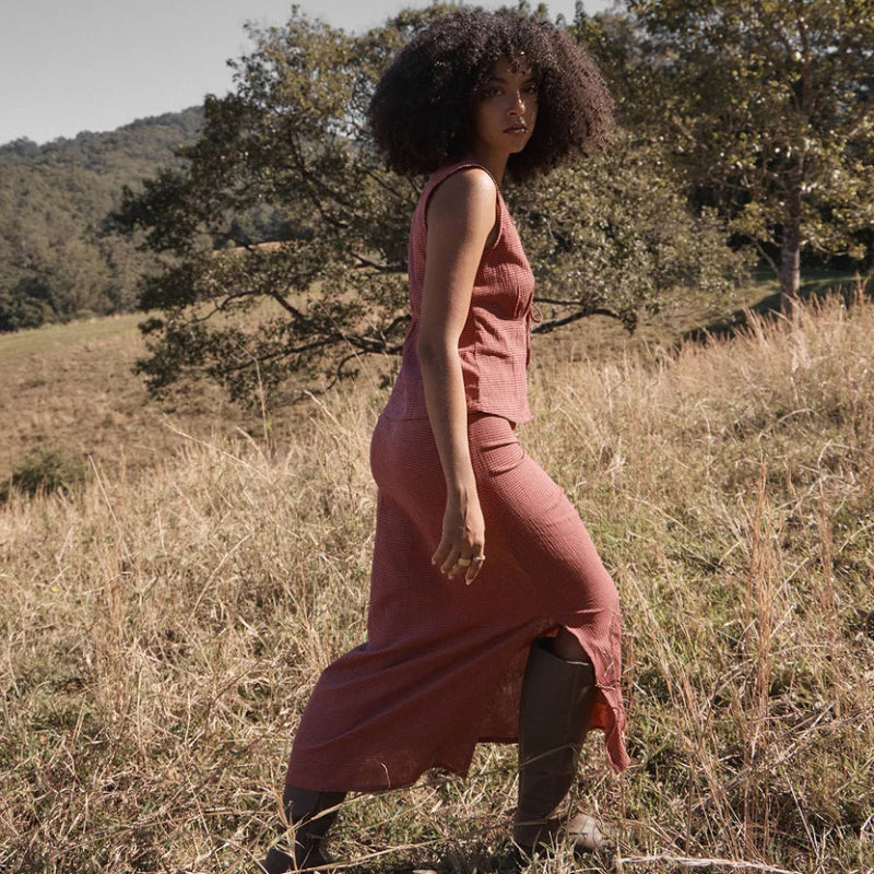 A woman with an afro walks through a dry grass field wearing a dusty rose sleeveless top  and matching skirt.