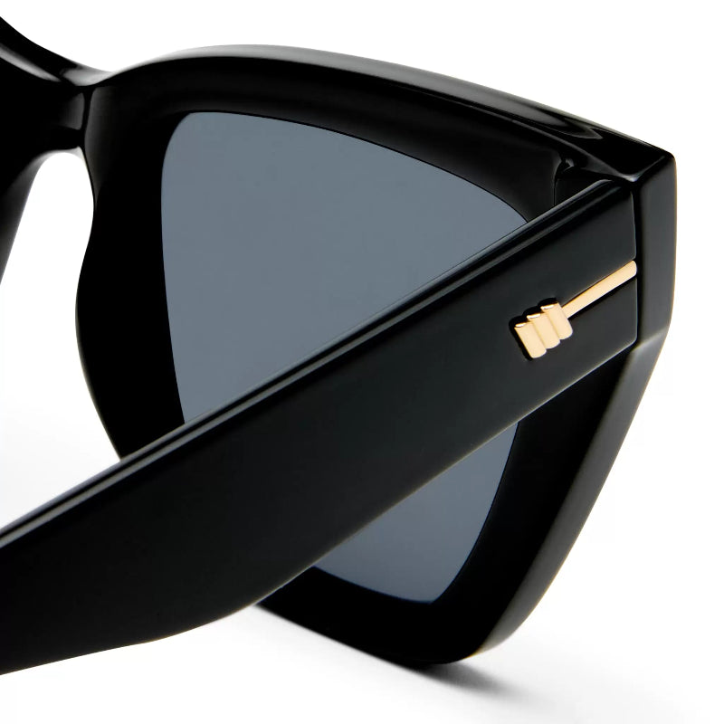 Close-up of black cat-eye sunglasses with dark gray lenses and a gold geometric accent on the temple arm, set against a clean white background.