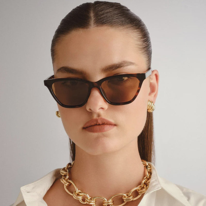 Woman wearing cat-eye sunglasses and a gold chain necklace against a neutral background