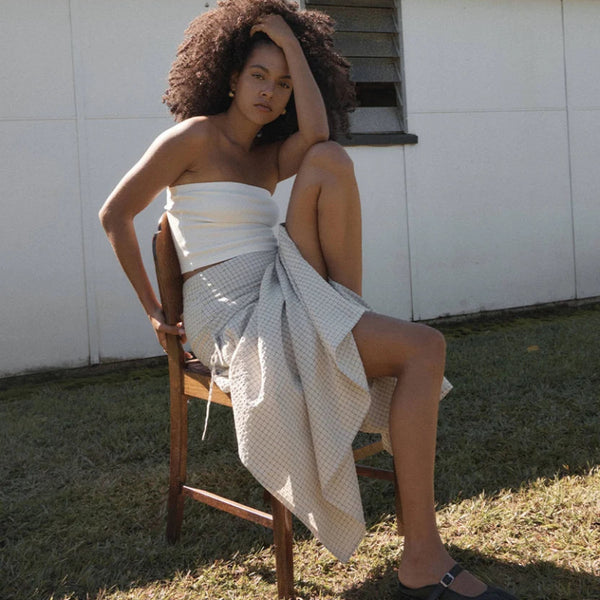 A woman with an afro sits on a wooden chair outdoors in front of a white building with a corrugated metal roof. She is wearing a white strapless top with a maxi skirt and dark flat shoes.