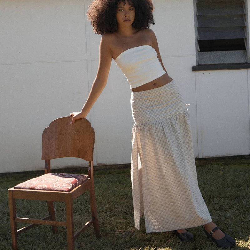 A woman stands outdoors on grass beside a wooden chair, wearing a white strapless bandeau-style top and a long cream textured skirt. A white building with a corrugated metal roof is behind her.