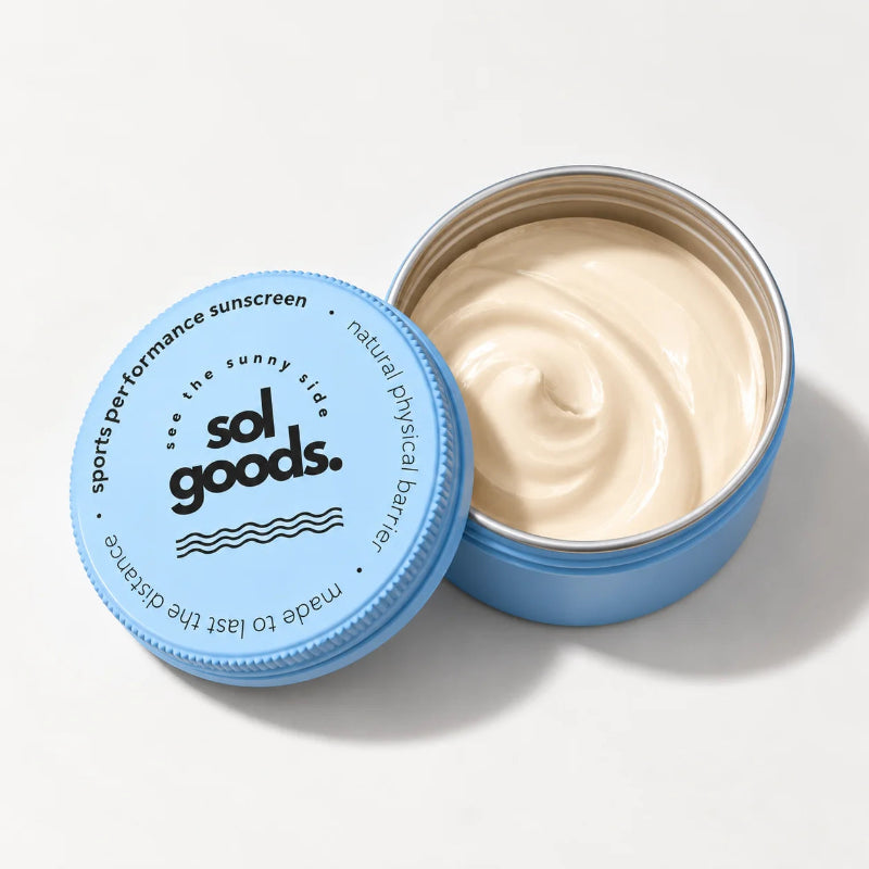 An open round blue tin of Sol Goods sports performance sunscreen, showing the cream-colored product inside, with the matching blue lid placed beside it on a clean white background.