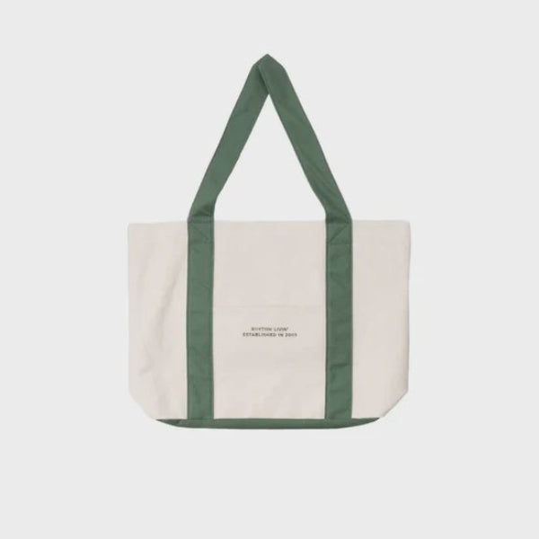 An off-white canvas tote bag with dark green handles and side trim, featuring a small printed logo on the front pocket, set against a light gray background.