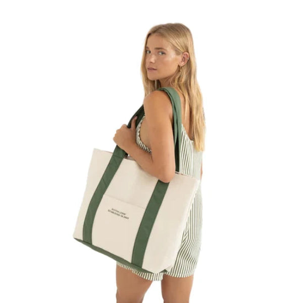 A woman wearing a green and white striped romper carries a large cream canvas tote bag with green shoulder straps against a plain white background.