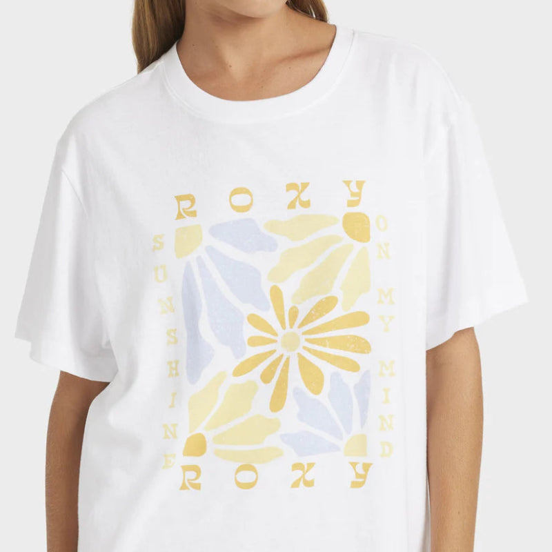 White t-shirt with yellow floral design and 'Roxy' branding worn by a person.
