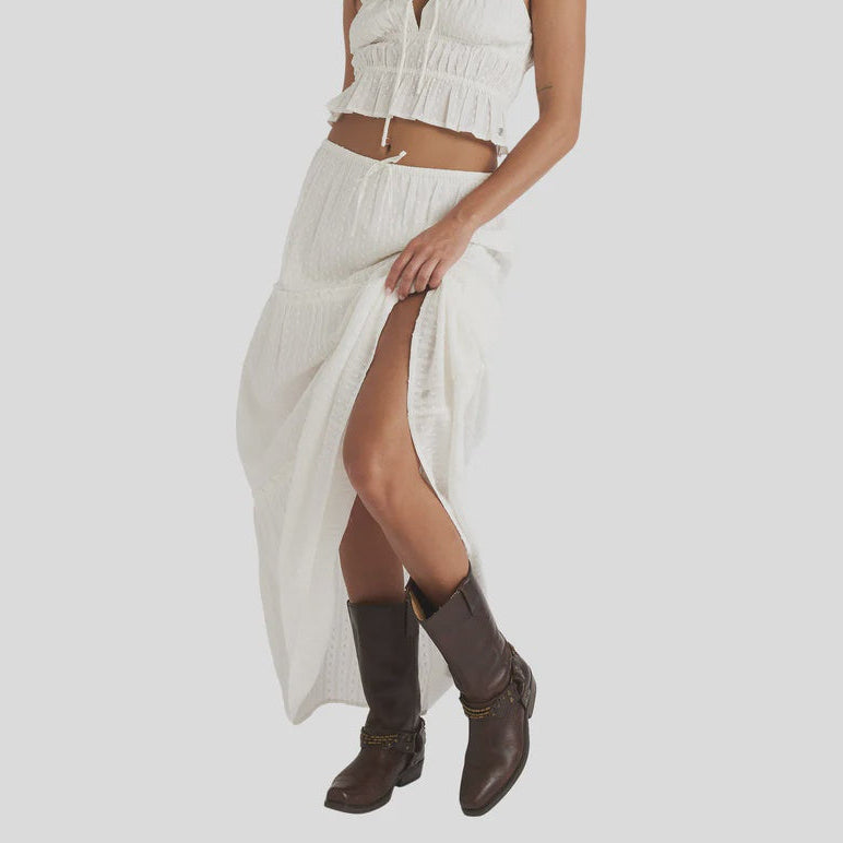 Woman wearing a white outfit with a crop top and long skirt, standing against a plain background.